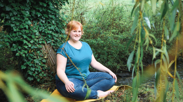 Fay, a white female with short, ginger hair, wearing dark blue jeans and a teal t-shirt sits with one knee bent and her arms around it. She is smiling to camera. She is sat on a yellow yoga mat in front of a tree covered in ivy