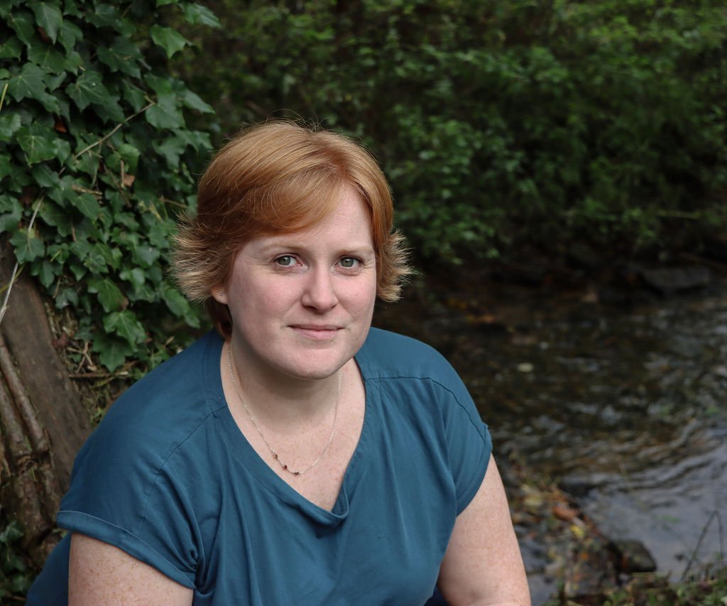 Fay, a white female with short, ginger hair, wearing a teal blue t-shirt, sits with her back against the trunk of a tree. She is looking up at the camera and there is a stream in the background